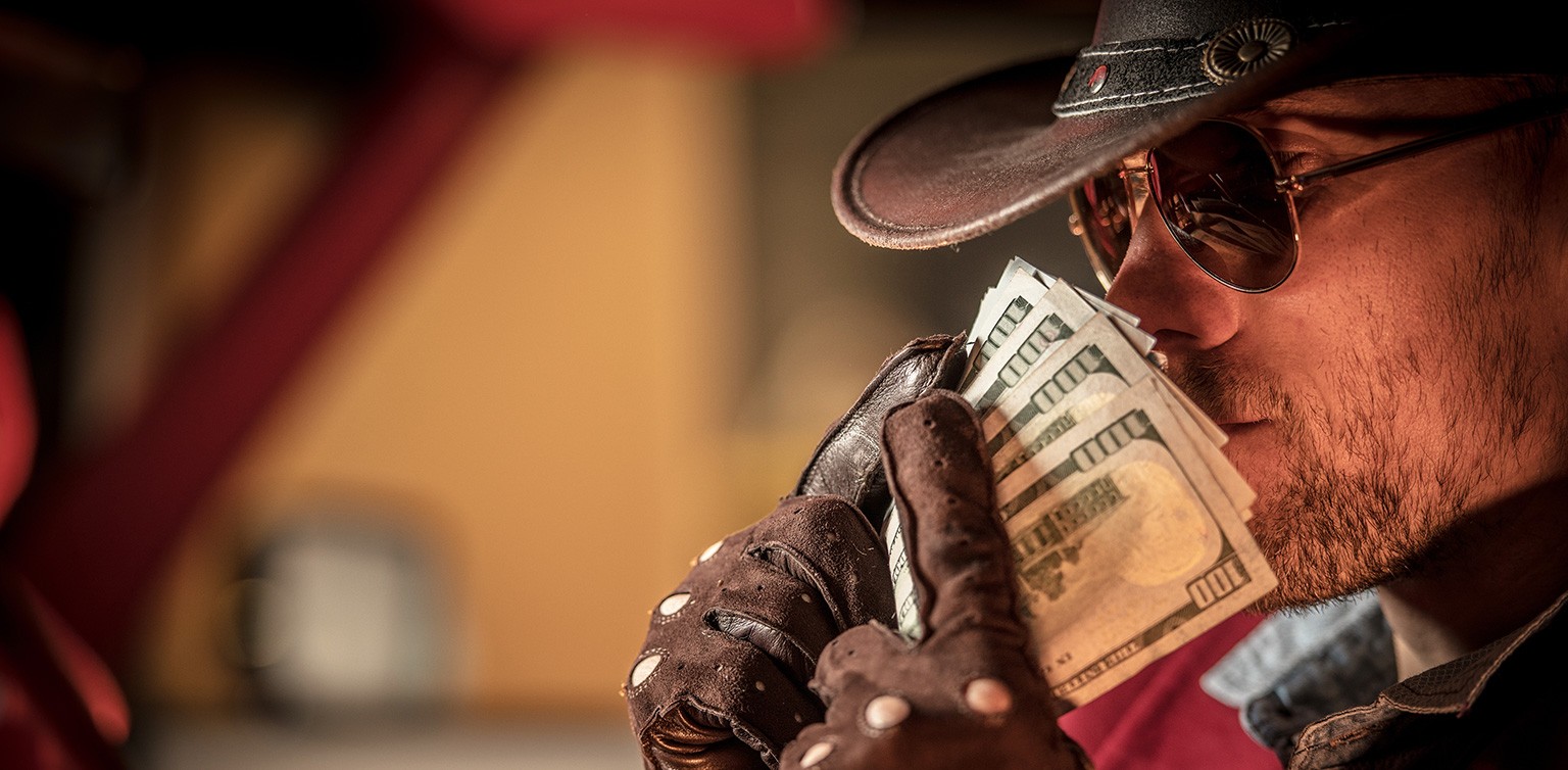 A photo of a cowboy wearing bare knuckle leather gloves and sunglasses holding up a wad of dollar bills to his nose